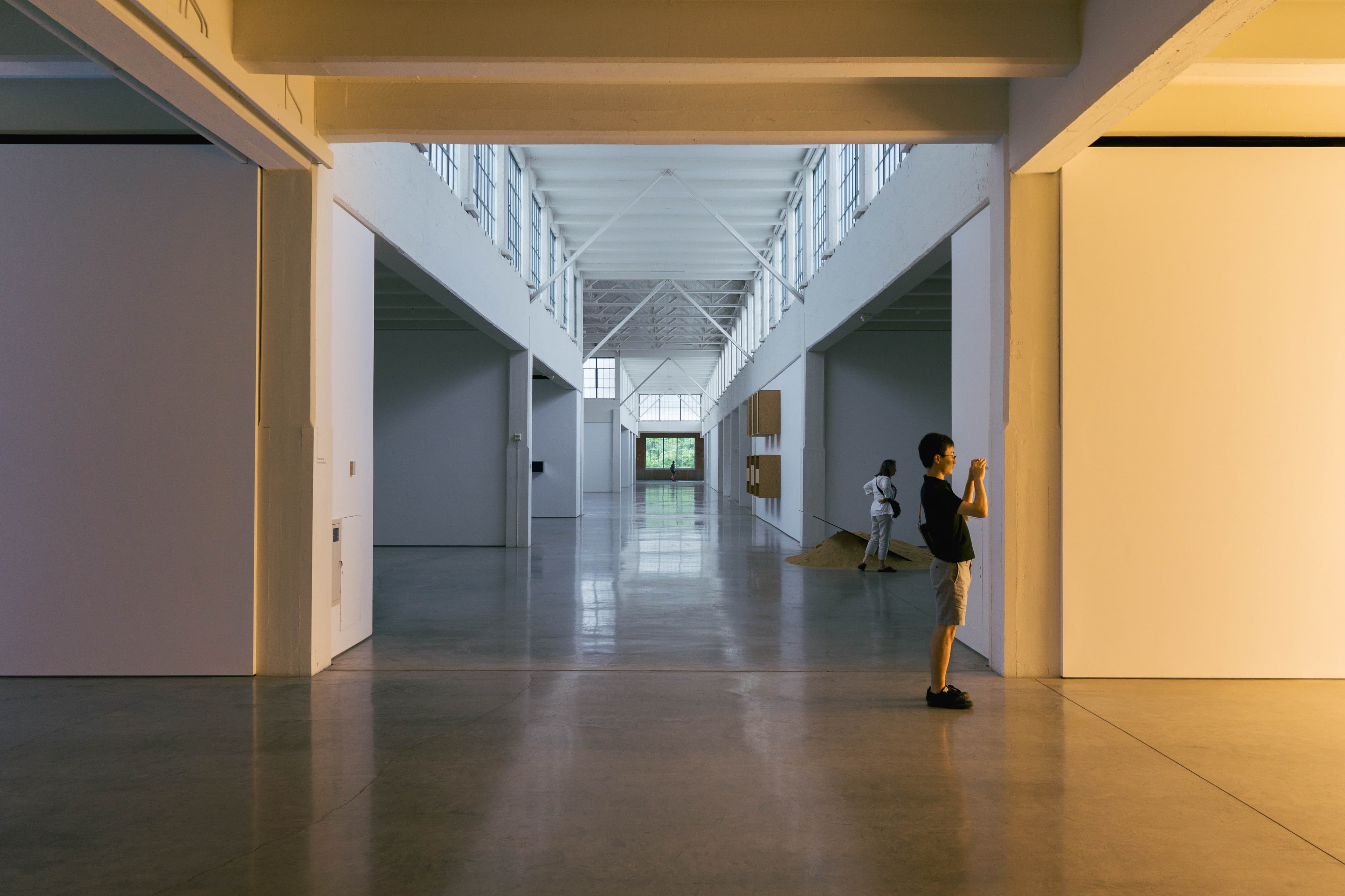 A man standing in a warmly-lit room and taking a photograph of something out of frame. Behind him is a long, daylight-drenched and spacious corridor. Close by, a woman stares at something else out of frame, and in the very far distance, green trees can be seen through a frosted window.