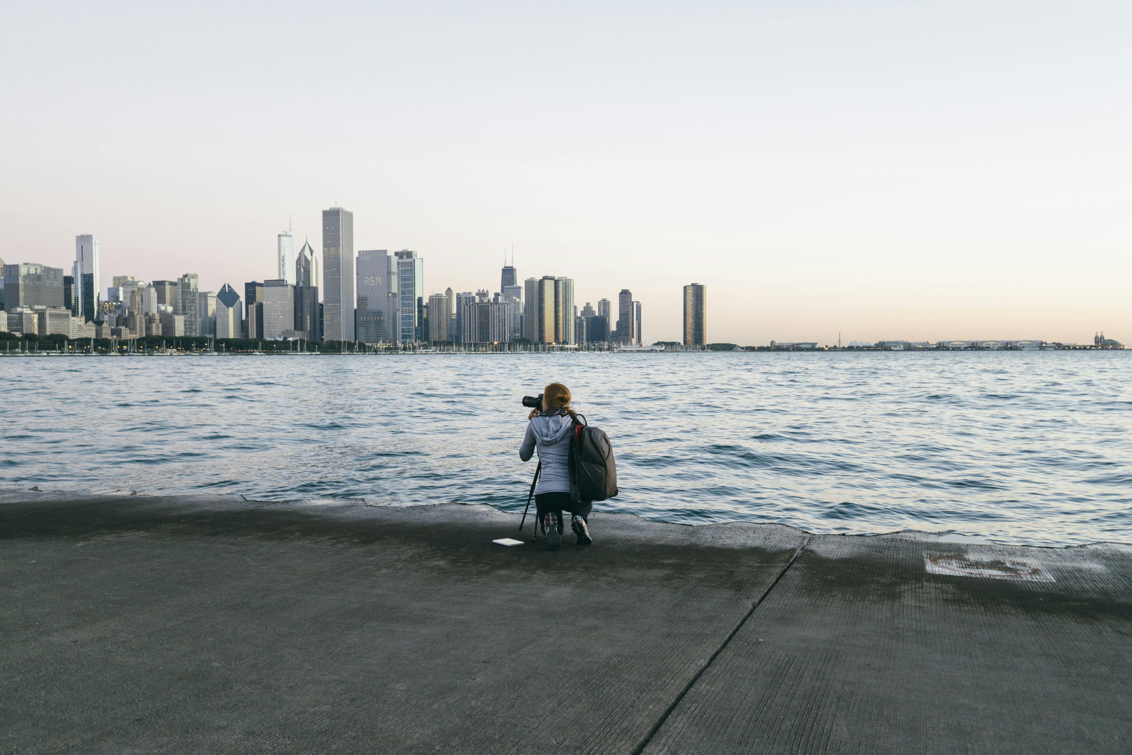 A woman crouches near a body of water at sunrise to take a photograph of a city in the distance.